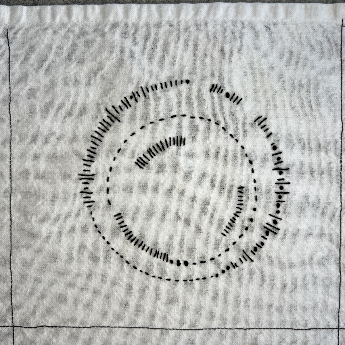 Overhead photo of white fabric 8”x8” square, with black embroidered lines. A illustration of a ribbon snaking up and down across the square. There are two sets of stepped light grey lines visible behind the ribbons.