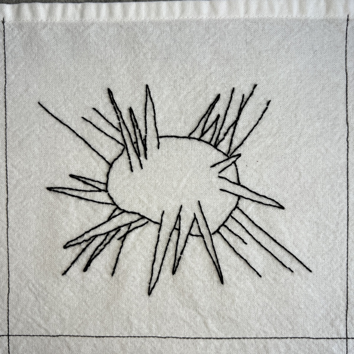 Overhead photo of white fabric 8”x8” square, with black embroidered lines. There’s a circular shape in the muffle of the square that has pokey and spiked lines coming out of it.