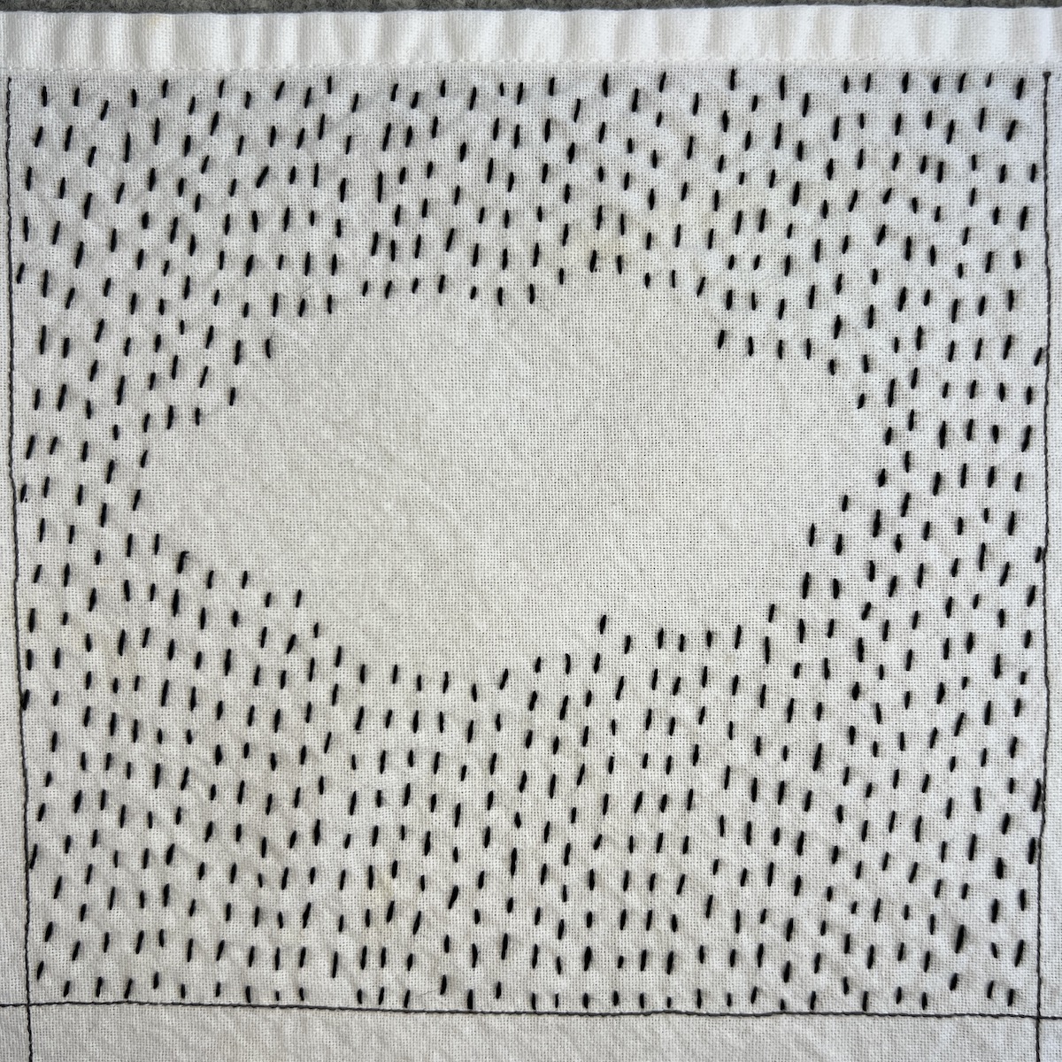 Overhead photo of white fabric 8”x8” square with rows of diagonal running stitch in black thread going across the square, except for negative space in the center in the shape of a cloud.