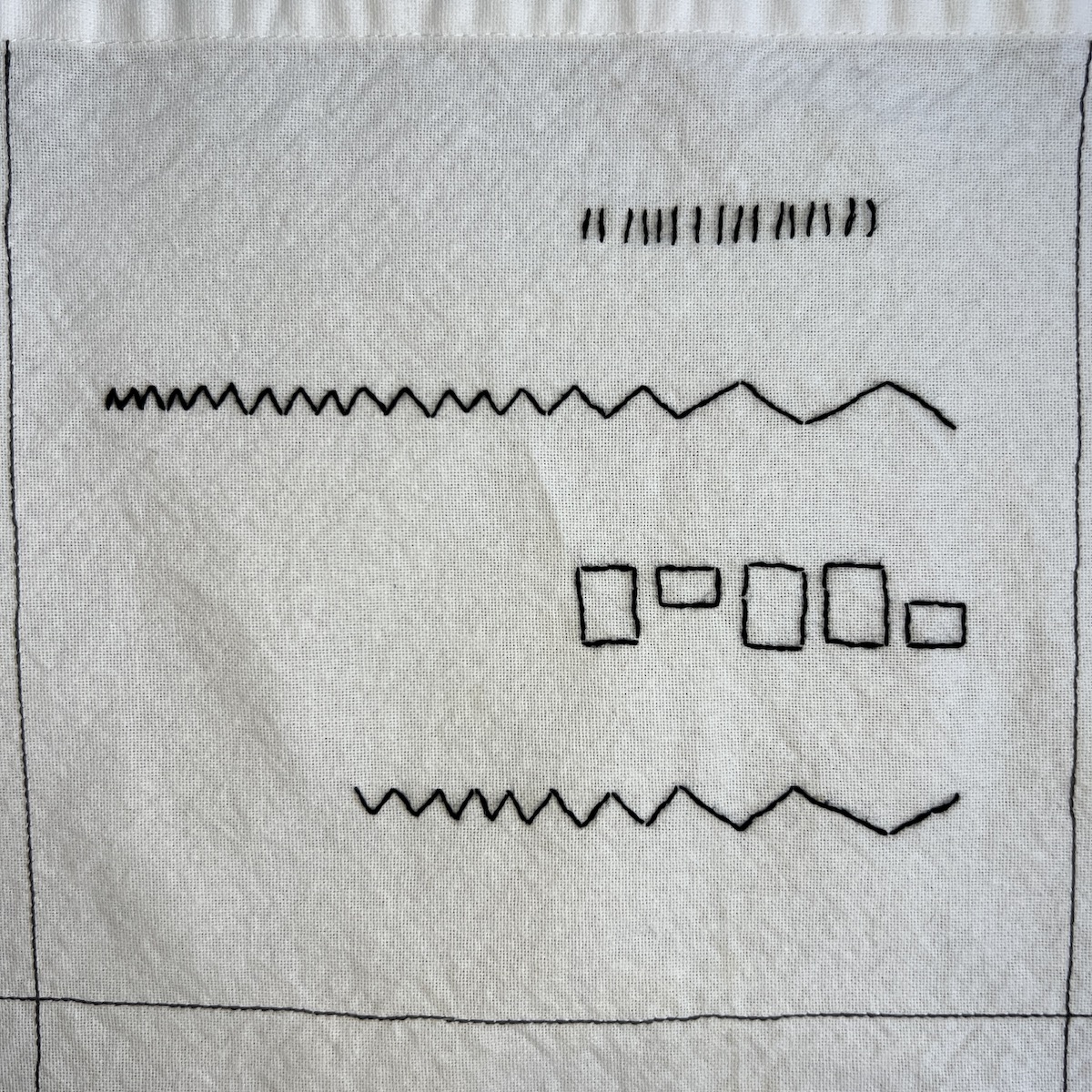 Overhead photo of white fabric 8”x8” square, with black embroidered lines. There are two horizontal jagged lines, one at the top going across the square, one at the bottom going from the midline to the the edge of the square. In between these two lines are outlines of five rectangles in a horizontal row. Above and to the right of the upper jagged line is a row of short vertical stitches.