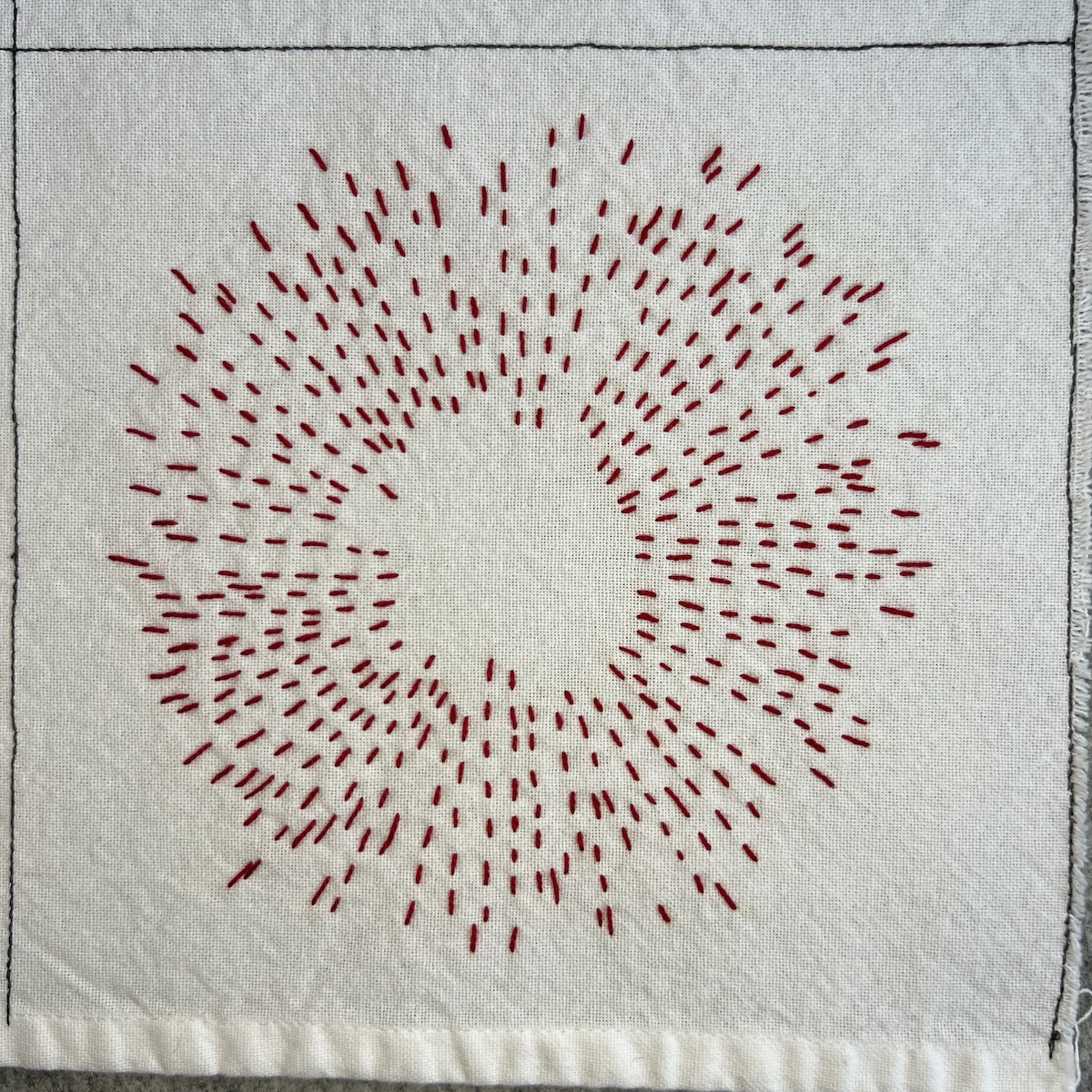 Overhead photo of white fabric 8”x8” square with a circle made of radiating running stitch lines embroidered in red thread.